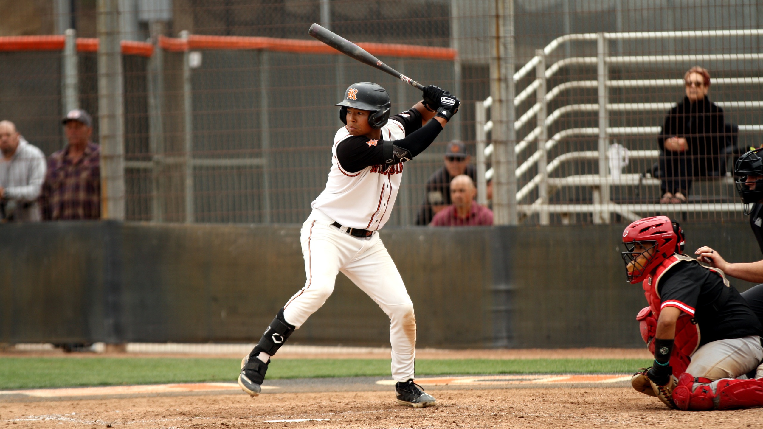 Baseball player at bat prepares to swing while the catcher and umpire are crouched behind home plate; spectators watch from the stands.