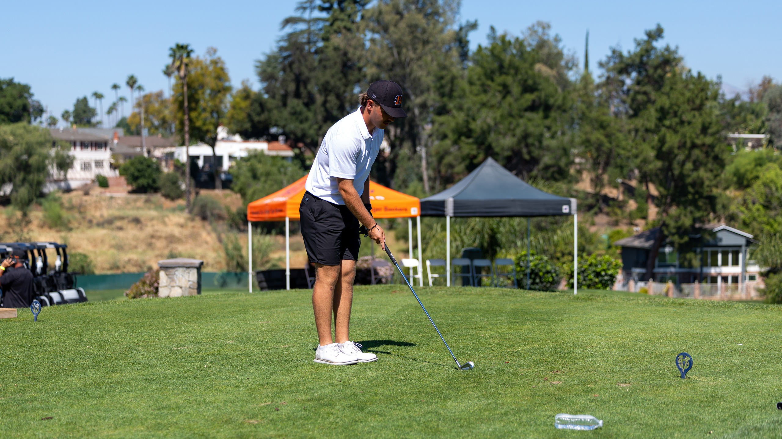 A man wearing a white shirt and black shorts prepares to hit a golf ball on the fairway.