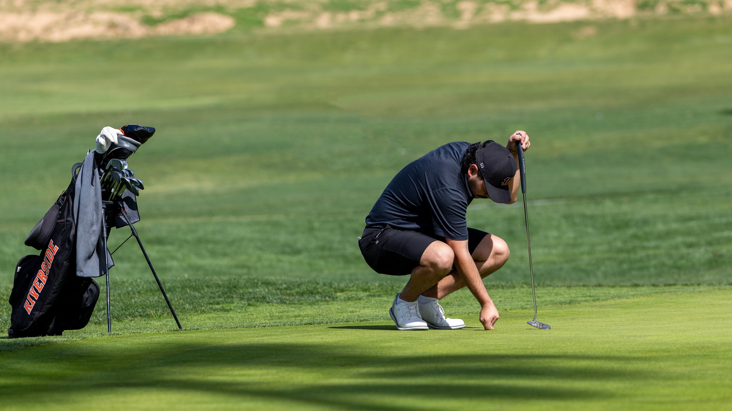 A golfer in black attire crouches on a green, focusing on the ball's position. A golf bag stands nearby. The scene is calm and focused.