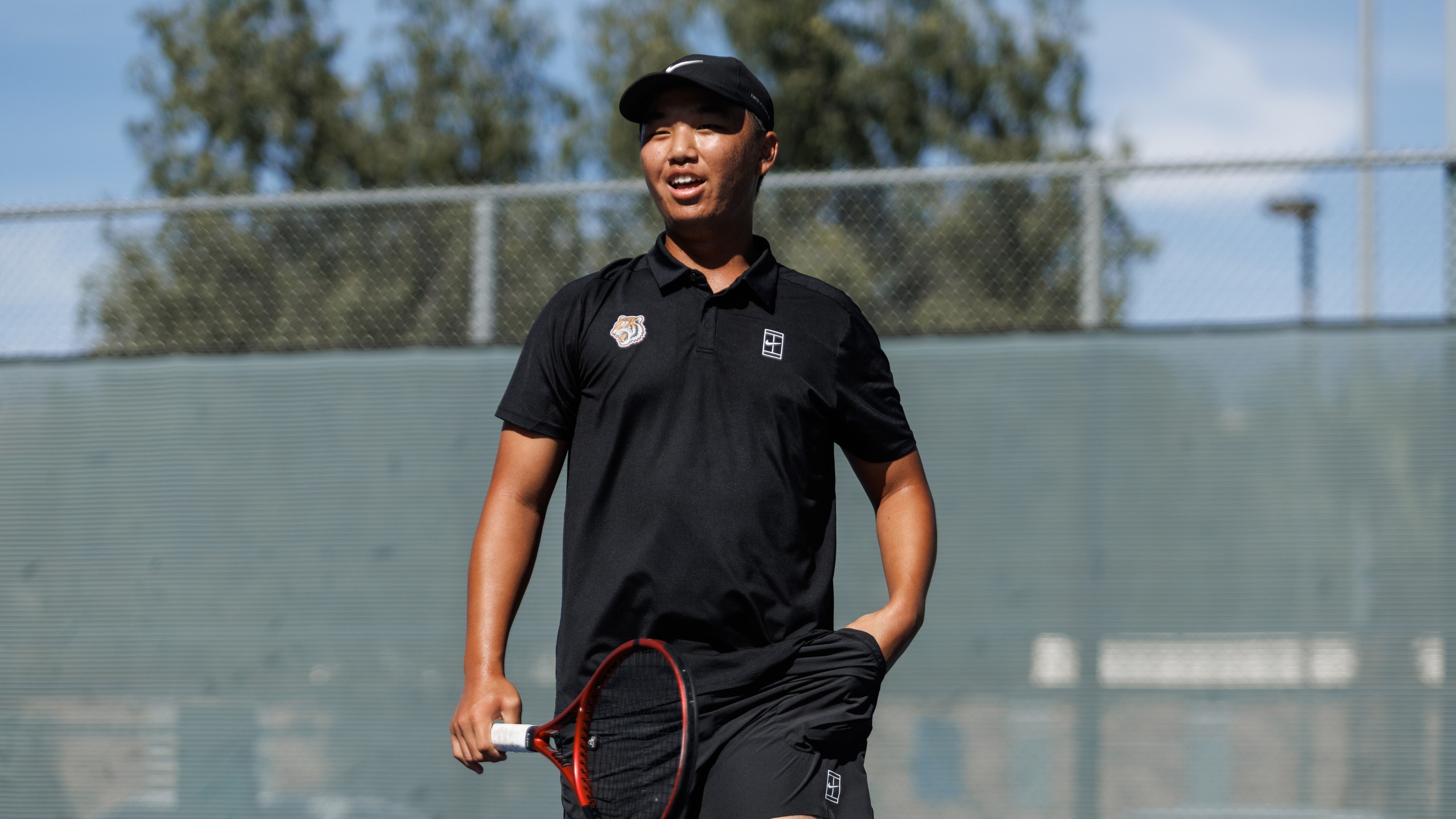 A man wearing a black sports outfit and cap holds a tennis racket, smiling brightly on a sunny day at the tennis court with trees in the background.