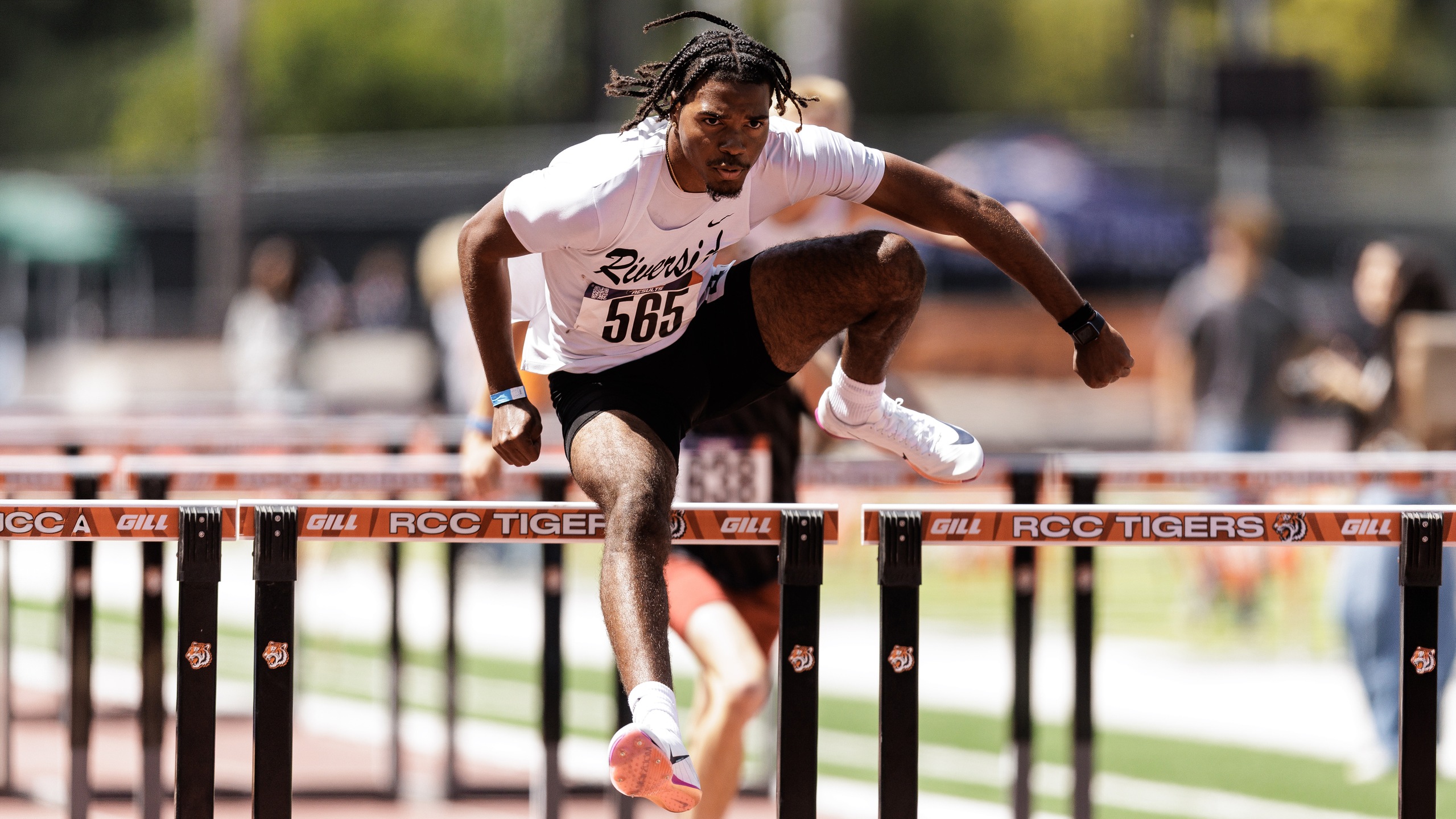 A men's track athlete in a white shirt and black shorts jumps over a hurdle.